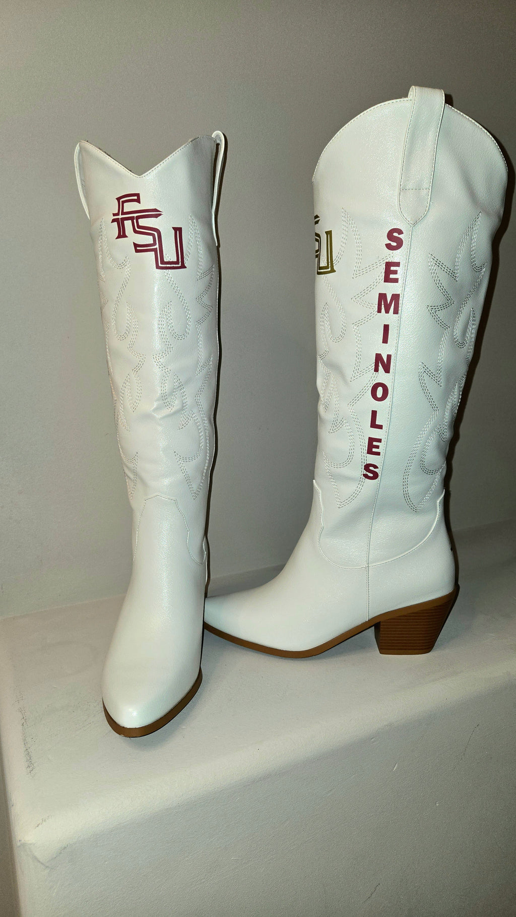 A pair of white faux leather cowboy boots featuring 'Seminoles' themed details, including vertical orange 'Seminoles' text on one boot and an orange and green 'U' logo on the other. Florida State Football Gameday