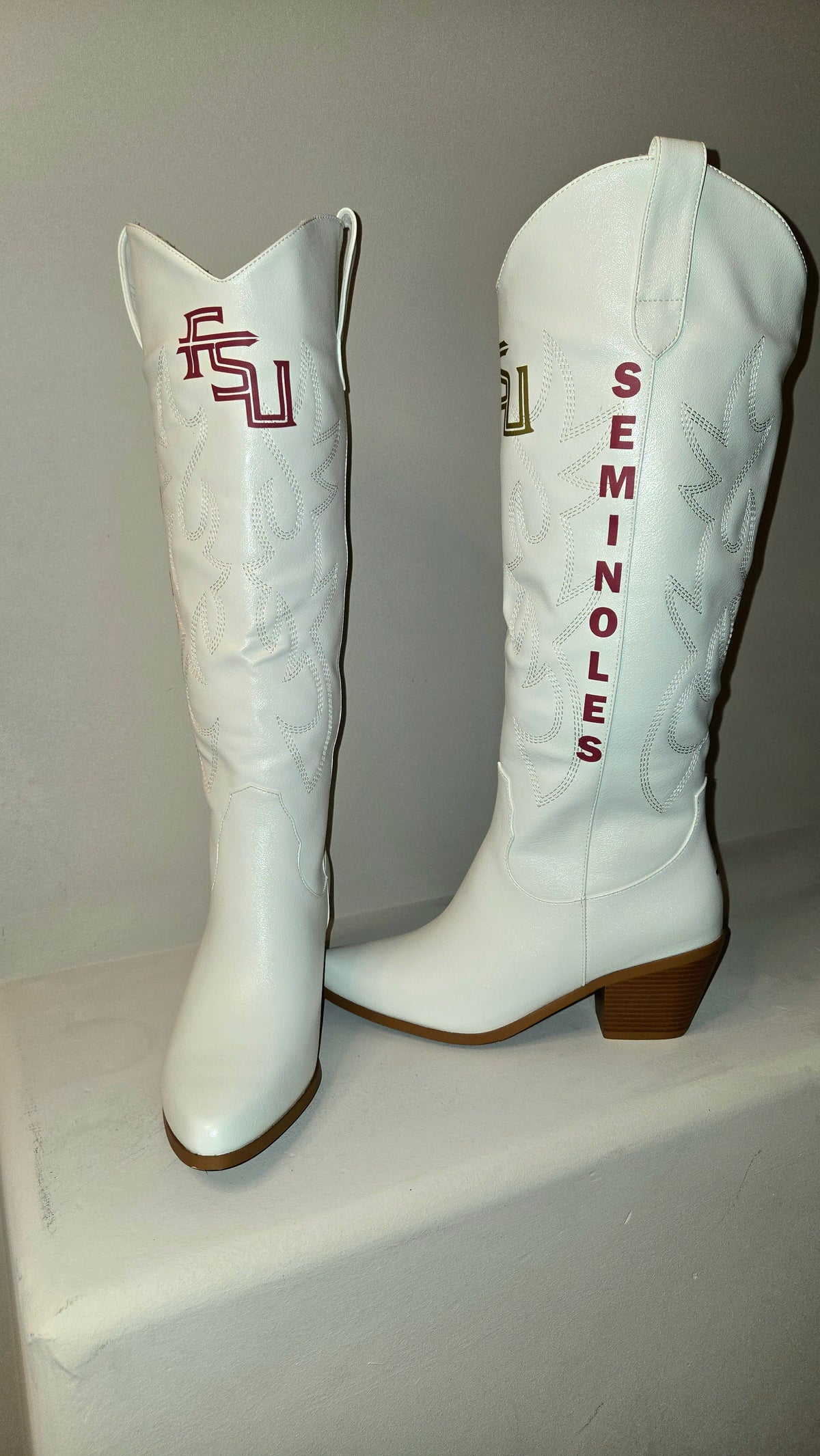 A pair of white faux leather cowboy boots featuring 'Seminoles' themed details, including vertical orange 'Seminoles' text on one boot and an orange and green 'U' logo on the other. Florida State Football Gameday